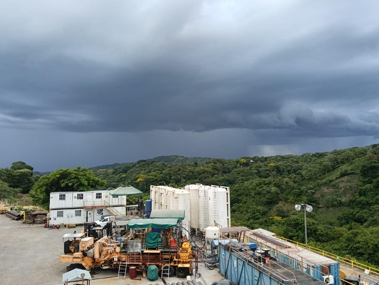 Well site with storage tanks and equipment below a green hillside under storm clouds