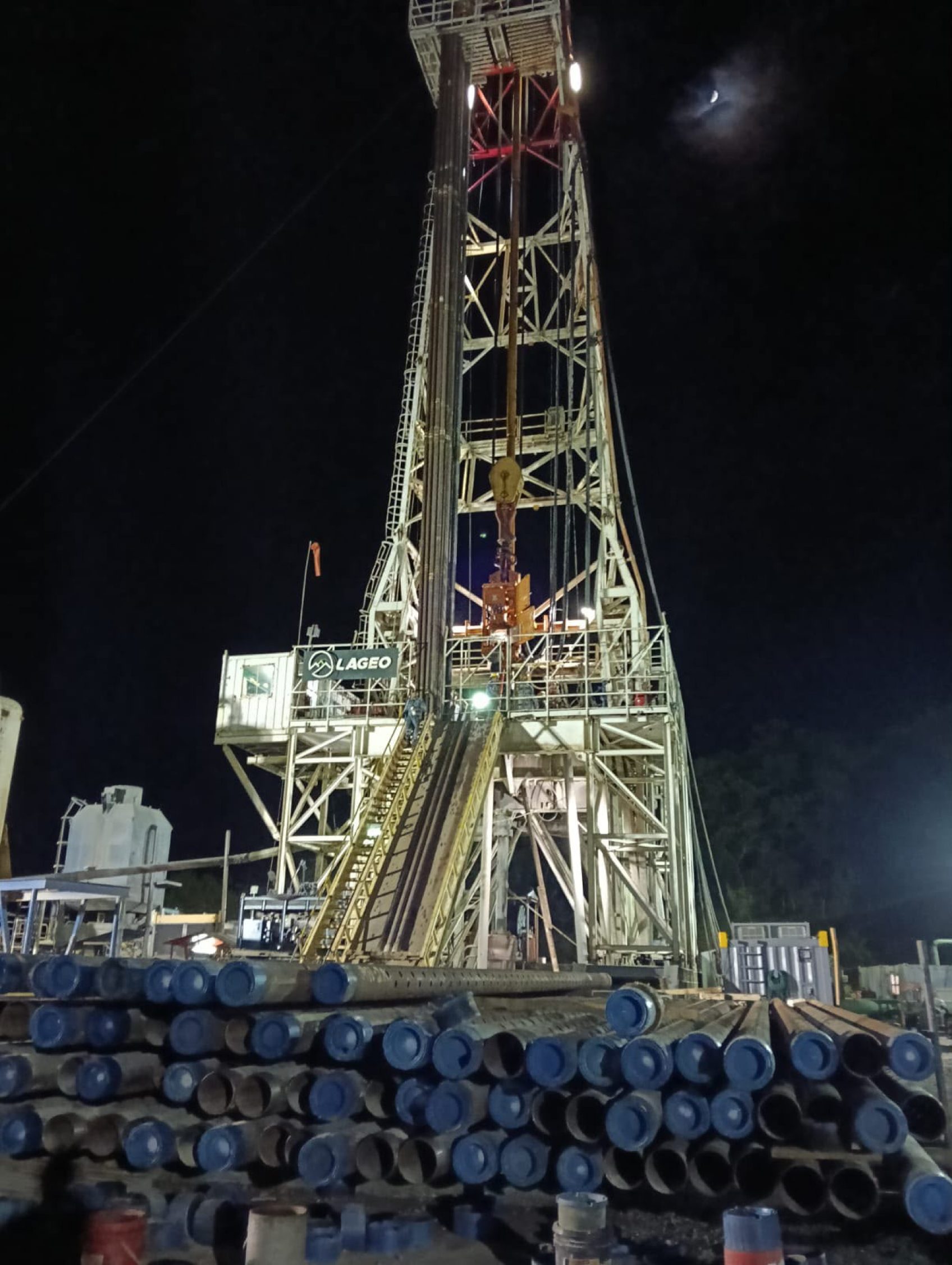 Drilling rig lit at night above stacked drill pipe